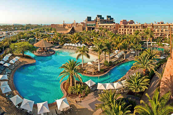 Et stort utendørsbasseng omgitt av parasoller, solsenger og grøntområder ved et feriehotell. (AI-generert tekst)