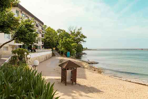 En bygning med flere balkonger ved siden av en sandstrand og hav med trær og en paviljong i forgrunnen. (AI-generert tekst)
