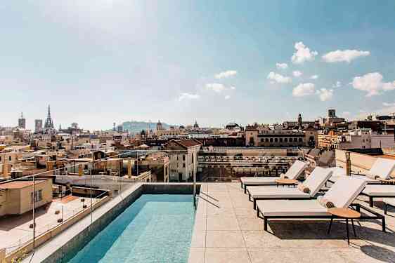 Et utendørsbasseng og solsenger på en takterrasse med utsikt over en by og dens bygninger under en klar himmel. (AI-generert tekst)