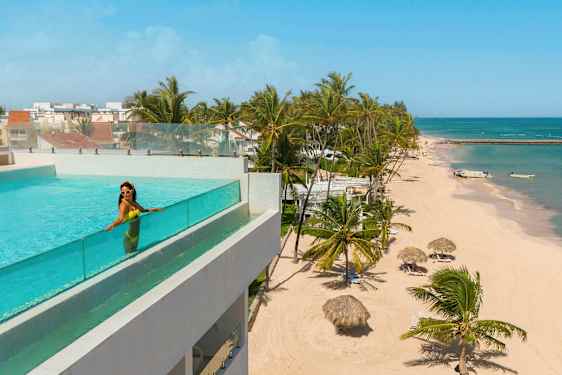 Et basseng med glassrekkverk på en bygning med utsikt over en strand med palmer og asurblått hav. (AI-generert tekst)