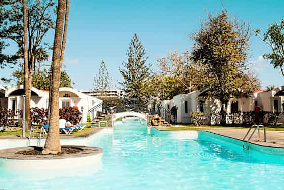 Et basseng med klart vann og en liten bro, omgitt av grøntområder og ferieleiligheter. (AI-generert tekst)