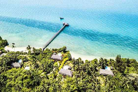 Flyfoto av en tropisk kystlinje med hus omgitt av grønnsaker. En brygge strekker seg ut i det turkise vannet. (AI-generert tekst)