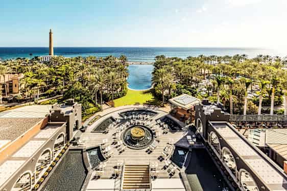 Utsikt over en hotellhage med palmer, et stort basseng og hav i bakgrunnen. (AI-generert tekst)