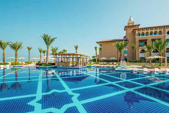 Et svømmebasseng med solsenger ved kanten, omgitt av palmer og en bygning i bakgrunnen. (AI-generert tekst)