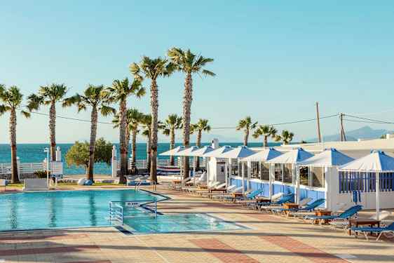 Et basseng med solsenger og parasoller under en klar himmel. Palmer og hav i bakgrunnen. (AI-generert tekst)