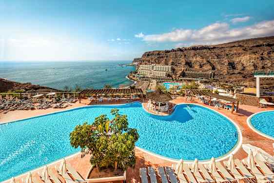 Hotellområde med bassenger, solsenger og utsikt over havet samt omkringliggende klipper. (AI-generert tekst)