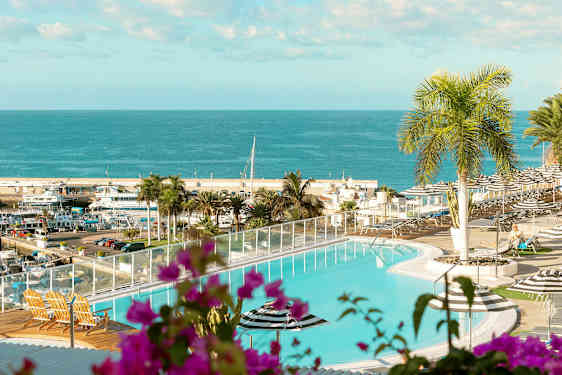 Et basseng med utsikt over et hav og en havn. I forgrunnen er planter og utemøbler. (AI-generert tekst)