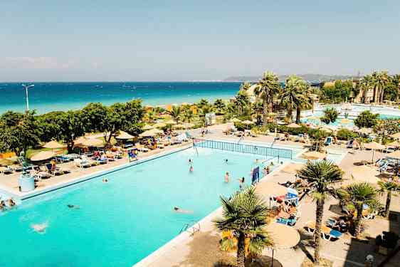 Et utendørsbasseng omgitt av solsenger og parasoller, med palmer og havet i bakgrunnen. (AI-generert tekst)