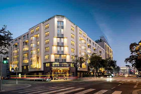 En bygning som er opplyst om kvelden, med flere etasjer og vinduer, plassert ved et veikryss. (AI-generert tekst)