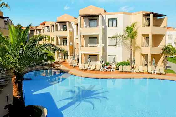 Et utendørsbasseng omgitt av solsenger og palmetrær ved en beige hotellbygning. (AI-generert tekst)