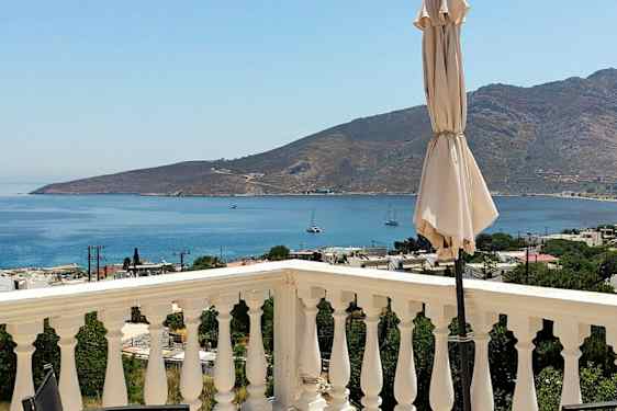 En utsikt fra en terrasse med rekkverk, en parasoll og havet og fjell i bakgrunnen. (AI-generert tekst)