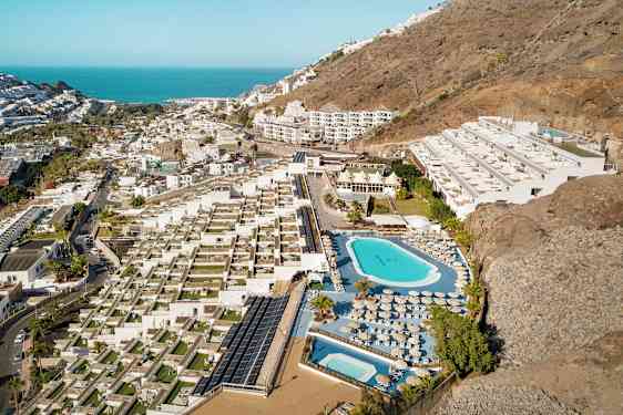 Flyfoto av et hotellområde med bassenger, solsenger og terrasser, plassert ved en skråning nær kysten. (AI-generert tekst)