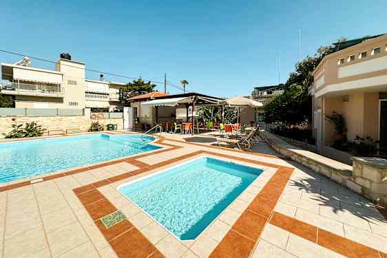 To utendørs bassenger omgitt av solsenger og et skyggeområde med fargerike stoler. (AI-generert tekst)