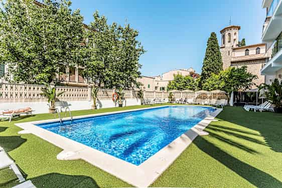 Utendørs svømmebasseng med solsenger rundt, omliggende landsbyarkitektur og grøntområder. (AI-generert tekst)