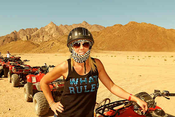 Kvinne står ved en rød firehjuling på et sandete ørkenområde med fjell i bakgrunnen. (AI-generert tekst)