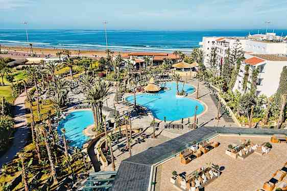 Hotellområde med bassenger og omkringliggende hager, plassert nær en sandstrand og havet. (AI-generert tekst)