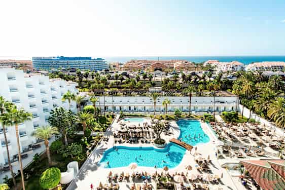 Utsikt over et hotellområde med bassenger, solsenger, palmer og bygninger i bakgrunnen. (AI-generert tekst)