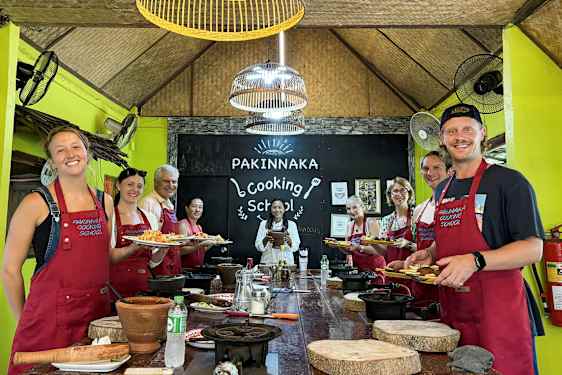 En gruppe personer med forklær står foran et skilt med teksten 'Pakinnaka Cooking School' i et matlagingsstudio. (AI-generert tekst)