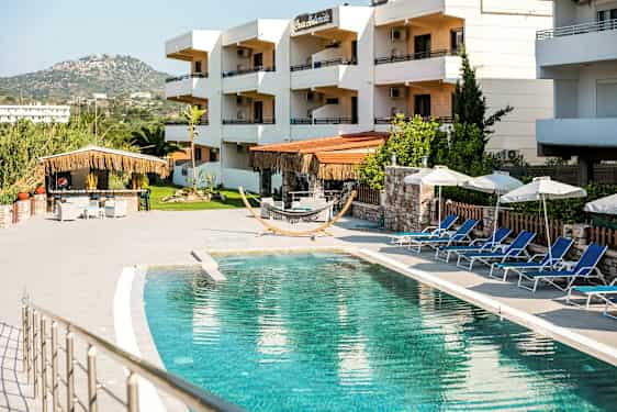 Utendørsbasseng med solsenger og parasoller, omgitt av bygninger i et ferieanlegg. (AI-generert tekst)