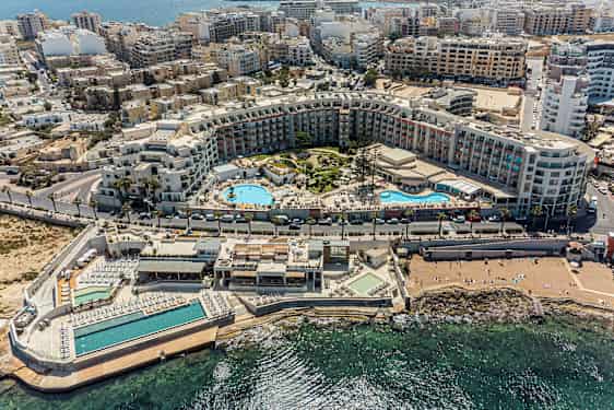 Bildet viser en omfattende ferieanlegg med bassenger, solområder og omkringliggende bygninger nær kysten. (AI-generert tekst)