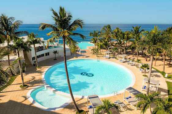 Et hotellområde med et stort rundt basseng omgitt av palmer, solsenger og cabanaer. Utsikt over havet i bakgrunnen. (AI-generert tekst)