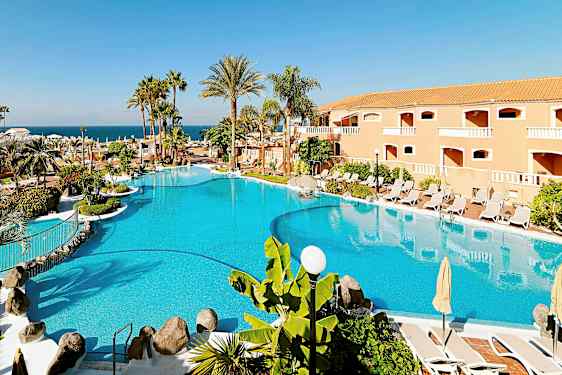 Et stort basseng omgitt av parasoller, solsenger, palmer og en bygning i bakgrunnen. (AI-generert tekst)