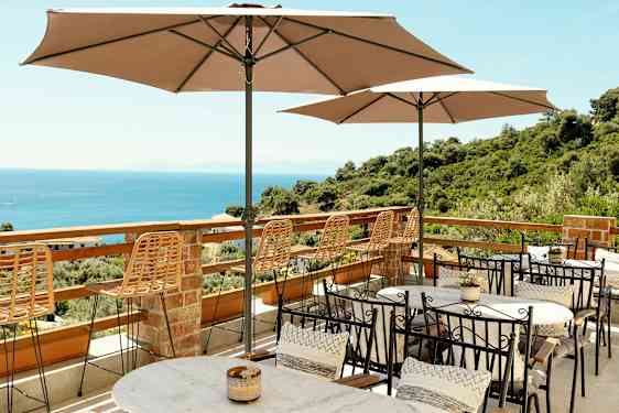 Terrasse med bord, stoler og parasoller med utsikt over havet og frodige omgivelser. (AI-generert tekst)