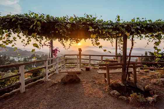 En utsiktspunkt med en trepergola dekket av blader samt benker og rekkverk rundt. Solnedgang over et fjellandskap i bakgrunnen. (AI-generert tekst)