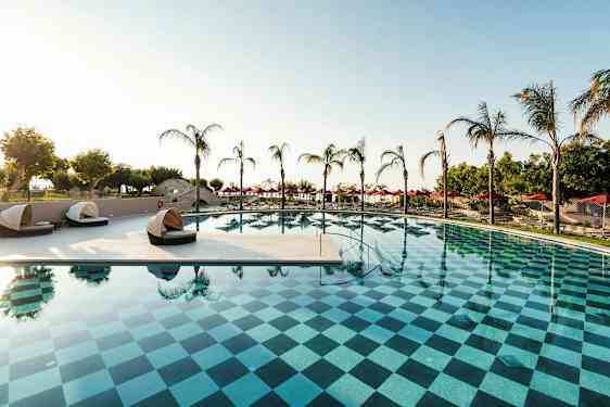 Et utendørsbasseng med rutemønster, omgitt av palmer og solsenger. (AI-generert tekst)