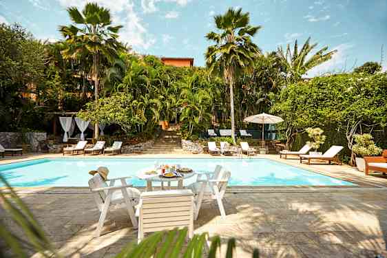 Et utendørs basseng omgitt av frodig vegetasjon og solstoler. Et bord med mat og drikke står ved bassengkanten. (AI-generert tekst)