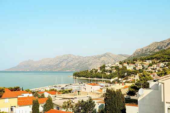 Utsikt over en kystby med røde tak, en småbåthavn og fjell i bakgrunnen. (AI-generert tekst)