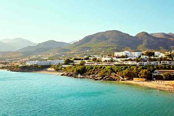 Flere bygninger nær en kyst med blått hav og fjell i bakgrunnen. (AI-generert tekst)