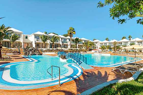 Et hotell med hvitmalte bygninger og et stort blått basseng foran, omgitt av palmer og grønne områder. (AI-generert tekst)