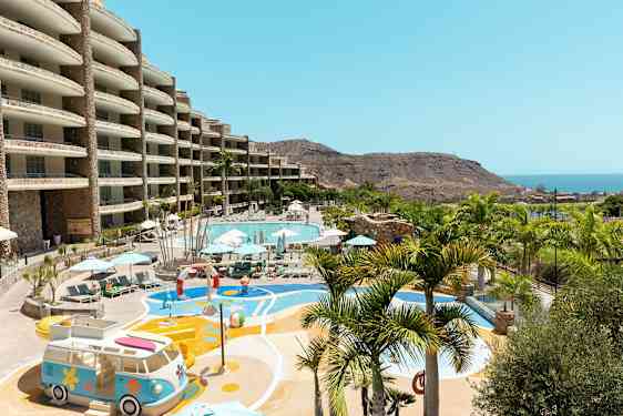 Utendørsområde med et stort basseng, omgitt av solsenger og parasoller, samt lekeområde med utsikt over havet og fjellandskap. (AI-generert tekst)