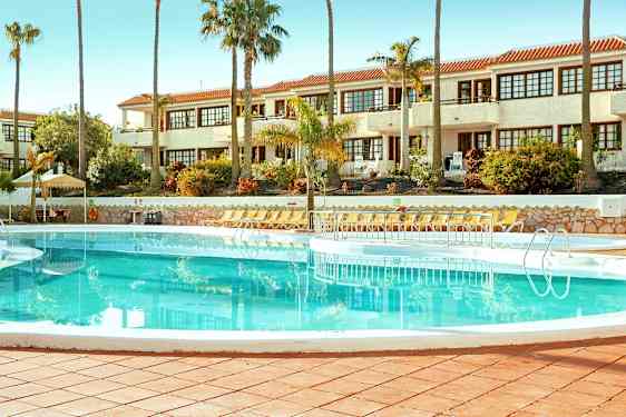 Et svømmebasseng omgitt av solsenger med en hvit bygning i bakgrunnen. (AI-generert tekst)