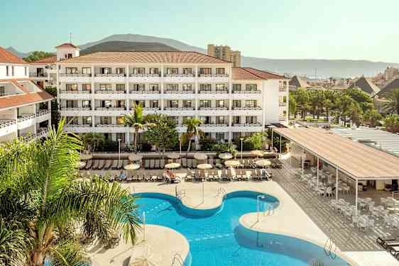 Hotellkompleks med basseng og solstoler, omgitt av grøntområder og fjell i bakgrunnen. (AI-generert tekst)