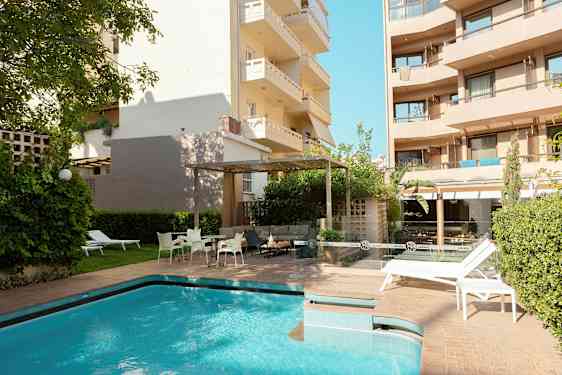 Utendørsområde med svømmebasseng og solsenger omgitt av bygninger og grøntområder. (AI-generert tekst)