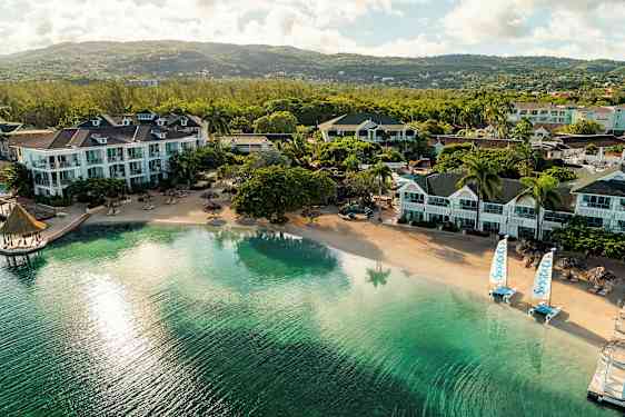 Flere hotellbygninger ved en rolig sandstrand med klart vann og utsikt over frodige åser. (AI-generert tekst)