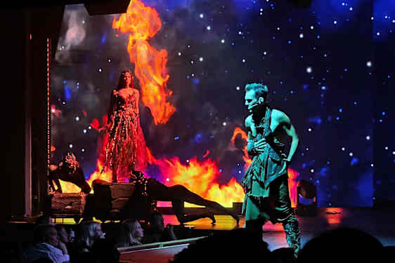 En scene med en kvinne i en ilddrakt omgitt av flammer og en mann i en svart drakt, med en stjernekledd bakgrunn. (AI-generert tekst)