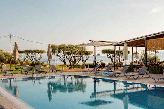 Utsikt over et basseng med solsenger rundt omkring, trær i bakgrunnen og havet i det fjerne. (AI-generert tekst)