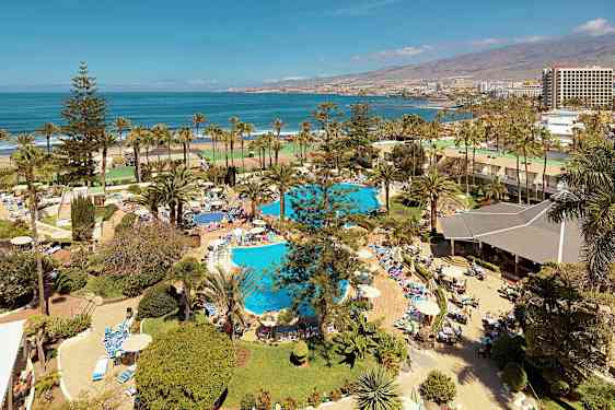 Utsikt over et feriested med bassenger, palmer og mennesker som soler seg og bader nær havet. (AI-generert tekst)