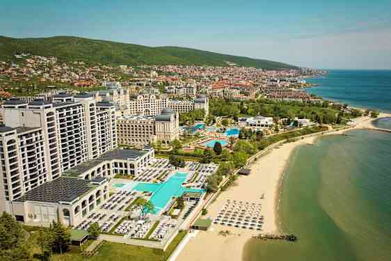 Hotell og bassenger ved stranden med havet og byen i bakgrunnen. (AI-generert tekst)