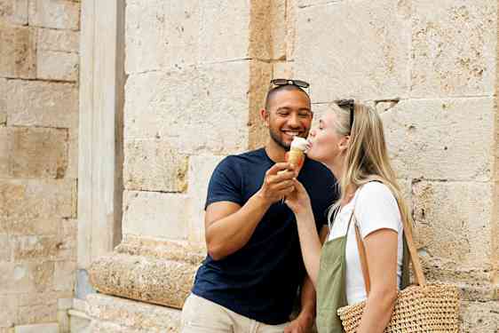 To personer står ved en steinbygning, hvor en av dem har en stråveske. (AI-generert tekst)