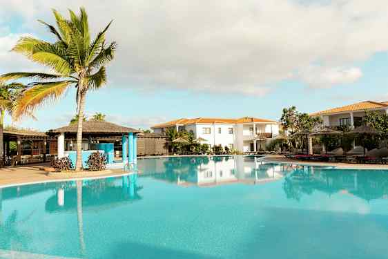 En tropisk bassengområde med palmer, parasoller og omkringliggende bygninger. (AI-generert tekst)