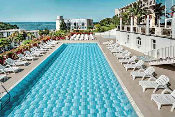 Et rektangulært basseng omgitt av solsenger, med havet og en bygning i bakgrunnen. (AI-generert tekst)