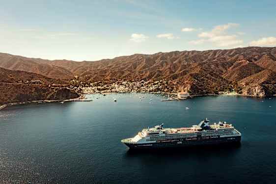 Et stort cruiseskip ligger i en bukt nær en by omgitt av fjell. (AI-generert tekst)