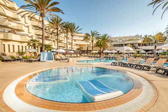 Et bassengområde på et hotell med solsenger og parasoller rundt bassenget, omgitt av palmer og balkonger. (AI-generert tekst)