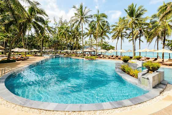 Et utendørs basseng omgitt av solsenger og parasoller ved en tropisk resort. (AI-generert tekst)