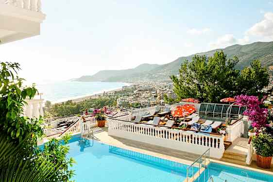 Utsikt over et basseng med solsenger, havet og en kystby i bakgrunnen. (AI-generert tekst)