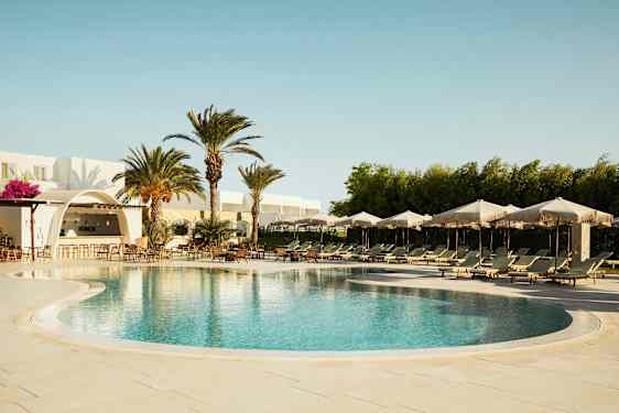 Et utendørs basseng med solsenger og parasoller, omgitt av palmer og en bygning med en bar. (AI-generert tekst)